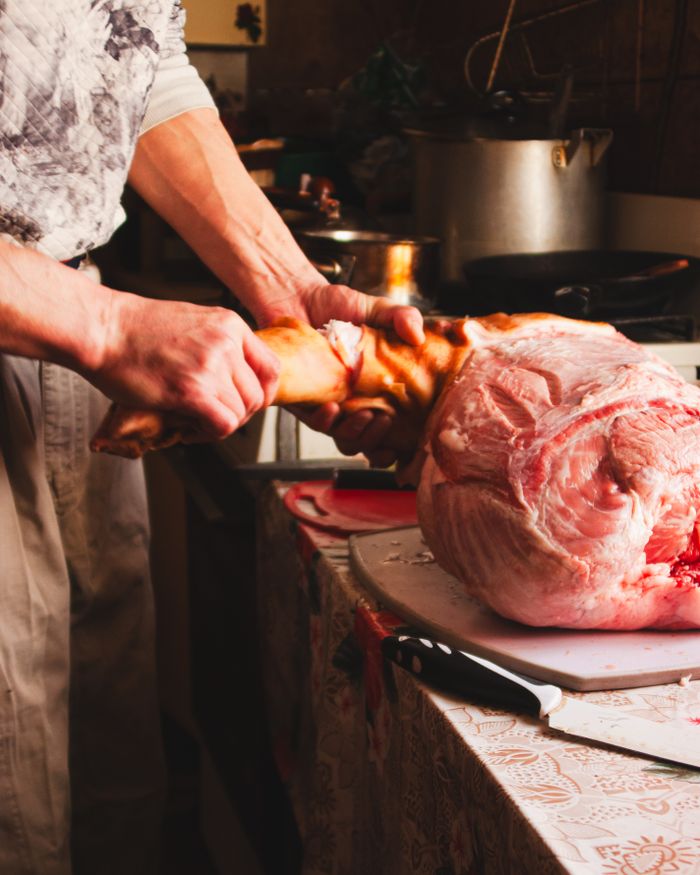 about-bg Chef skillfully butchering a raw pork leg in a kitchen using sharp cutlery.