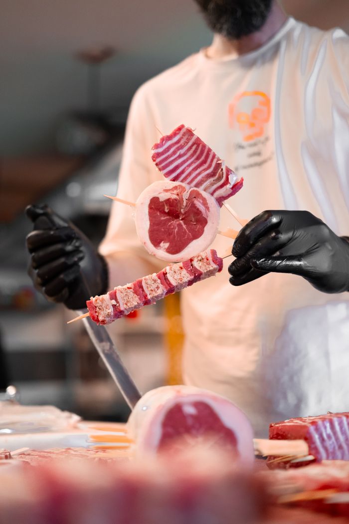 our-story A butcher skillfully prepares skewered red meat in an indoor setting, showcasing culinary expertise.